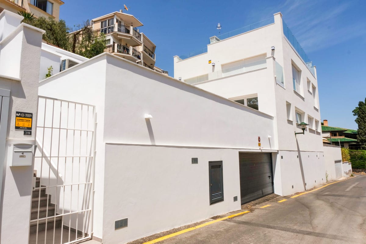 MONTE SANCHA 28 photographed from the street: a white façade with a white metal entrance gate, stairs, dark garage door and several storeys visible beneath a blue sky.