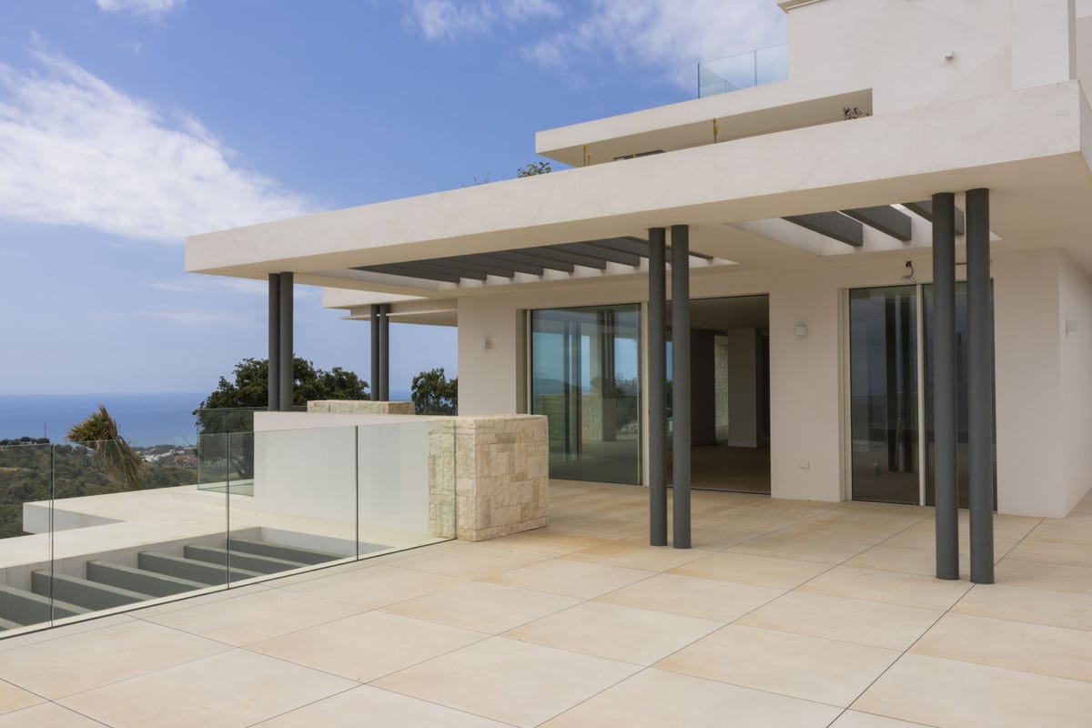 OCEAN VIEW MARBELLA: spacious beige tiled terrace with a concrete pergola, dark round columns, two wide sliding doors to the interior, a glass balustrade with a travertine block feature, steps down to a lower level and the sea visible in the distance.