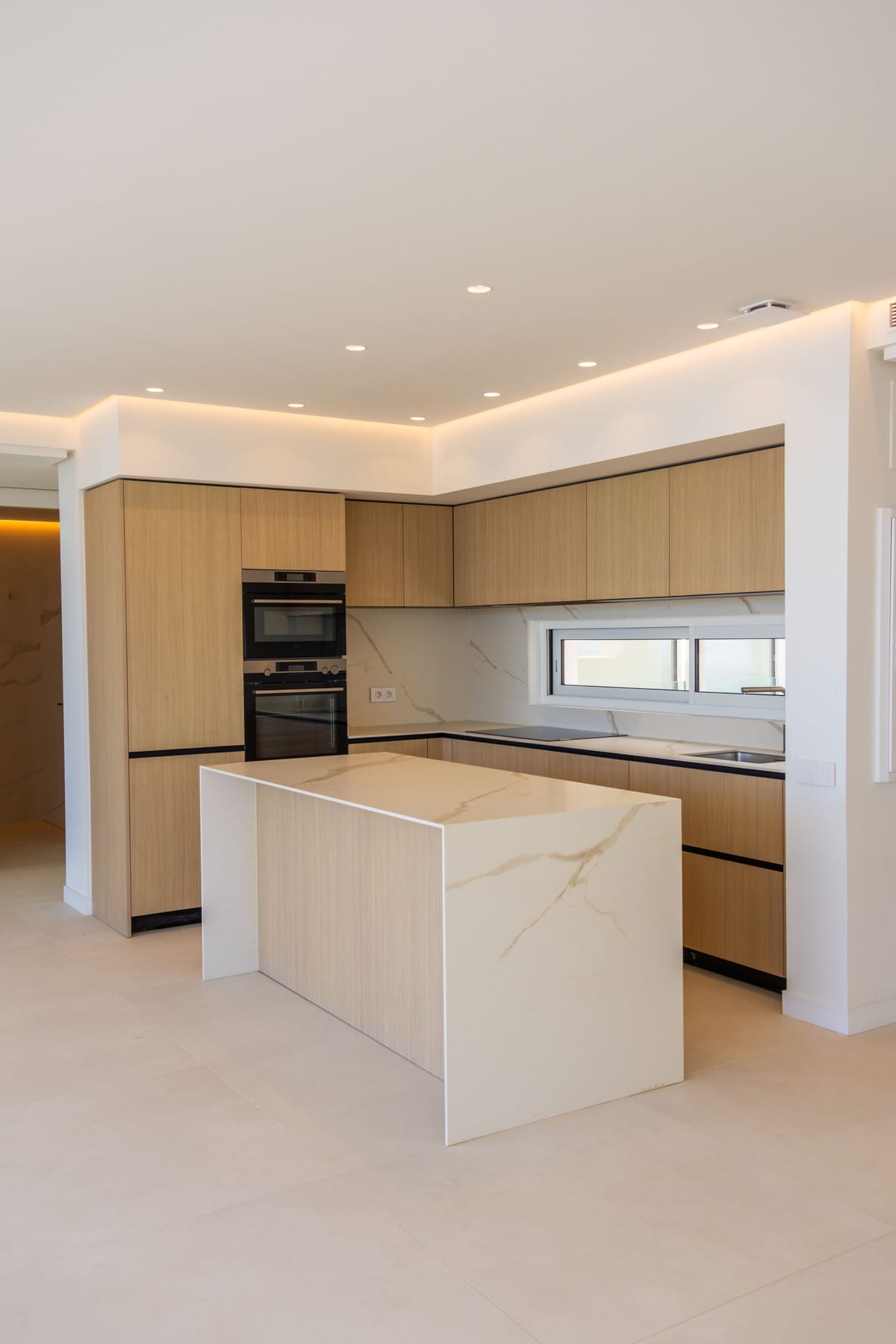 OCEAN VIEW MARBELLA: kitchen with light wood cabinetry, built-in double oven, white kitchen island with a gold-veined marble-effect worktop, light tiled floor and indirect ceiling lighting with recessed spotlights.