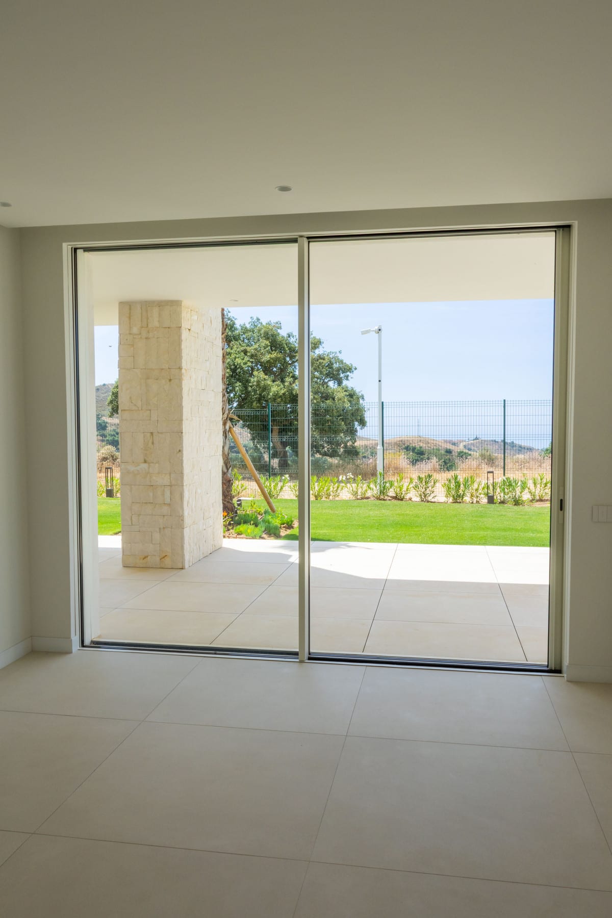 OCEAN VIEW MARBELLA: empty interior with a light tiled floor and large aluminium sliding door opening onto a tiled terrace with a natural stone column, green lawn, shrubs, green fencing and hilly landscape.