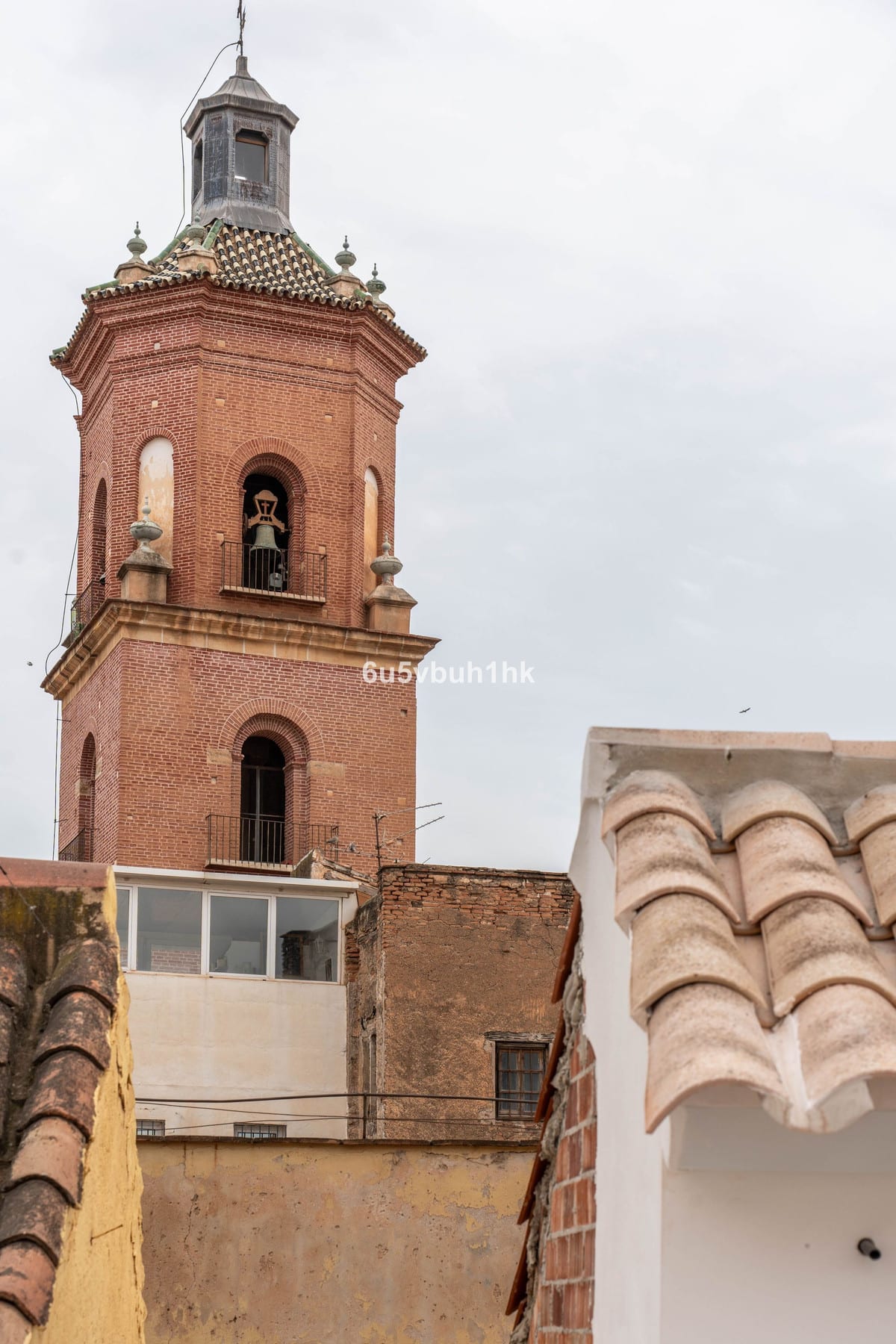 POZOS DULCES 29 — photograph of a brick bell tower with a visible bell, surrounded by tiled roofs and rendered façades beneath an overcast sky.