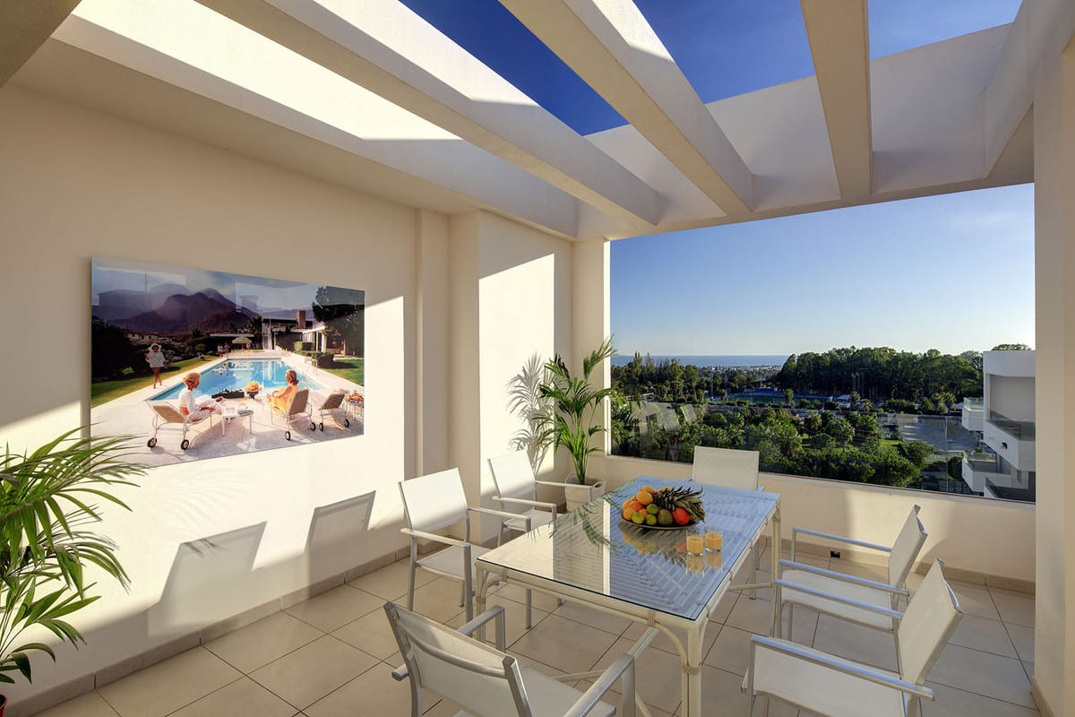 Project visualisation of a covered terrace at RESIDENCIAL ALBATROS XIV with a glass dining table, white chairs, exposed roof beams and views of greenery and a blue expanse of water on the horizon.