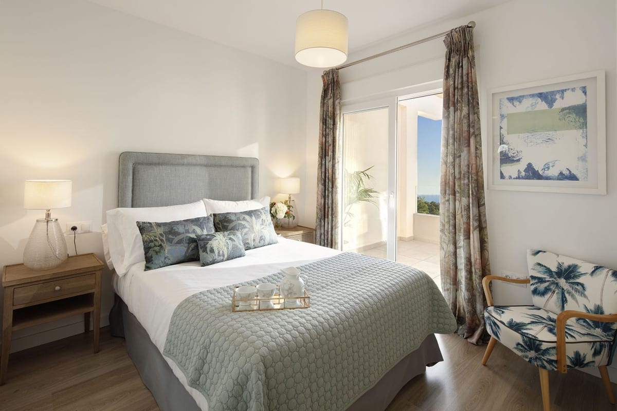 Bedroom at RESIDENCIAL ALBATROS XV with an upholstered grey bed, wooden bedside tables, armchair with palm-tree print and sliding glass door to a sunny terrace.