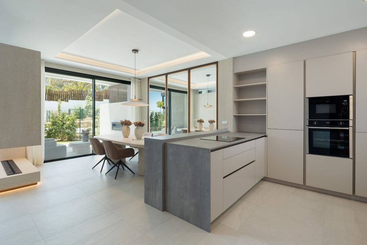 A dark grey concrete-effect kitchen island forms the centrepiece of the open-plan space, alongside tall cabinets with built-in appliances, a dining table and glazed sliding doors.