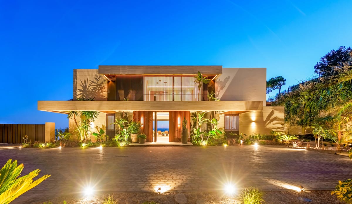 Entrance facade of THE SKY MARBELLA VILLAS at dusk, with a paved forecourt, ground lighting and planting around the central entrance.
