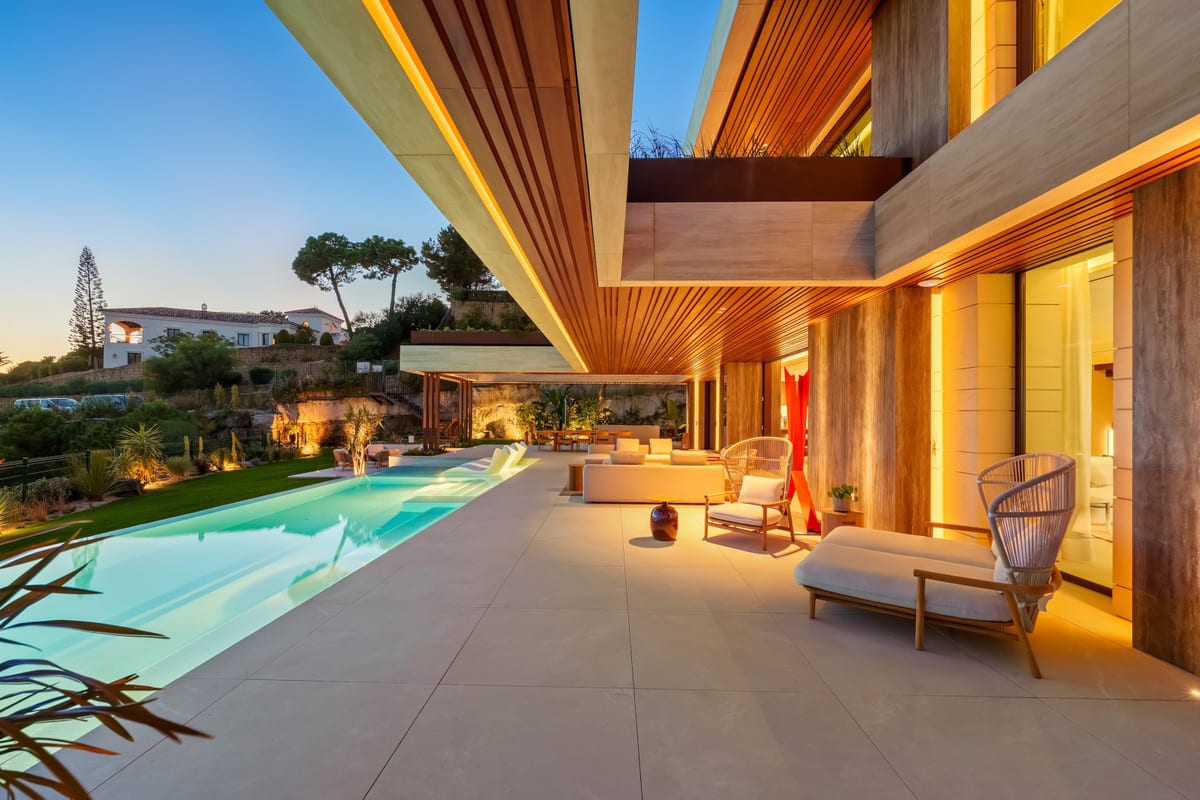 Covered terrace of THE SKY MARBELLA VILLAS at dusk, beside the illuminated pool, with a timber ceiling and lounge seating.