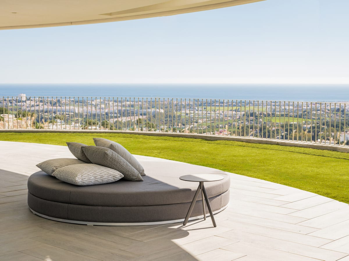 View of a covered terrace with a round daybed, a strip of lawn and the coastline in the distance.