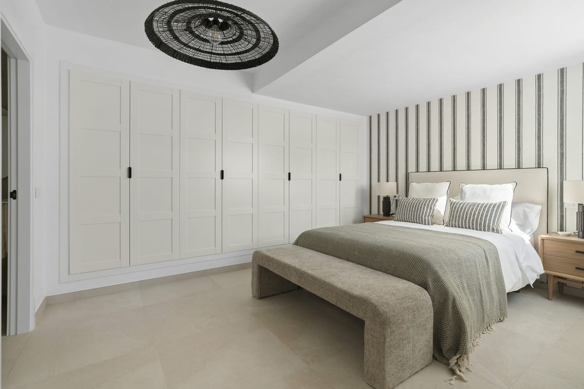 URBANIZACIÓN OLIMPO bedroom with a made-up double bed, striped wallpaper, an upholstered footstool, wooden bedside tables and a white fitted wardrobe wall.