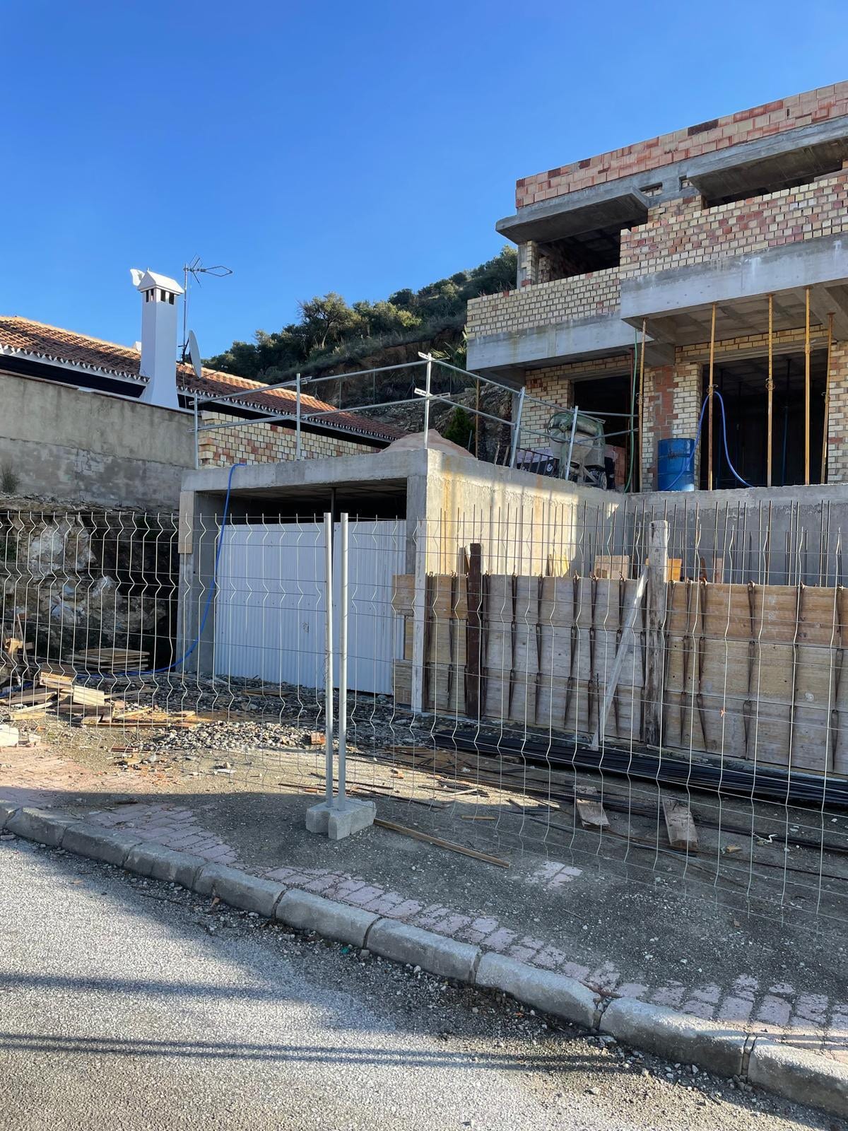 VILLA ALCALÁ DEL VALLE under construction, viewed from the street: brick façade walls across two storeys, concrete floor edges, timber formwork boards along the plot boundary and a temporary metal construction fence in the foreground.