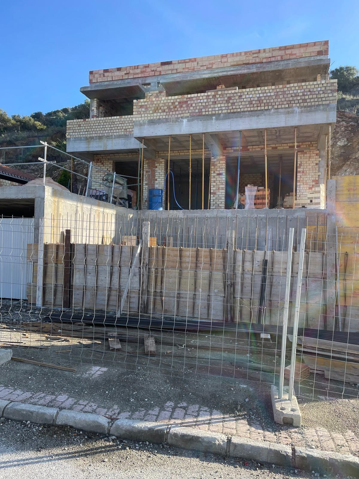 Front view of VILLA ALCALÁ DEL VALLE under construction: a brick façade across two storeys with open façade openings, vertical reinforcement bars, timber formwork boards along the plot boundary and a metal construction fence in the foreground.