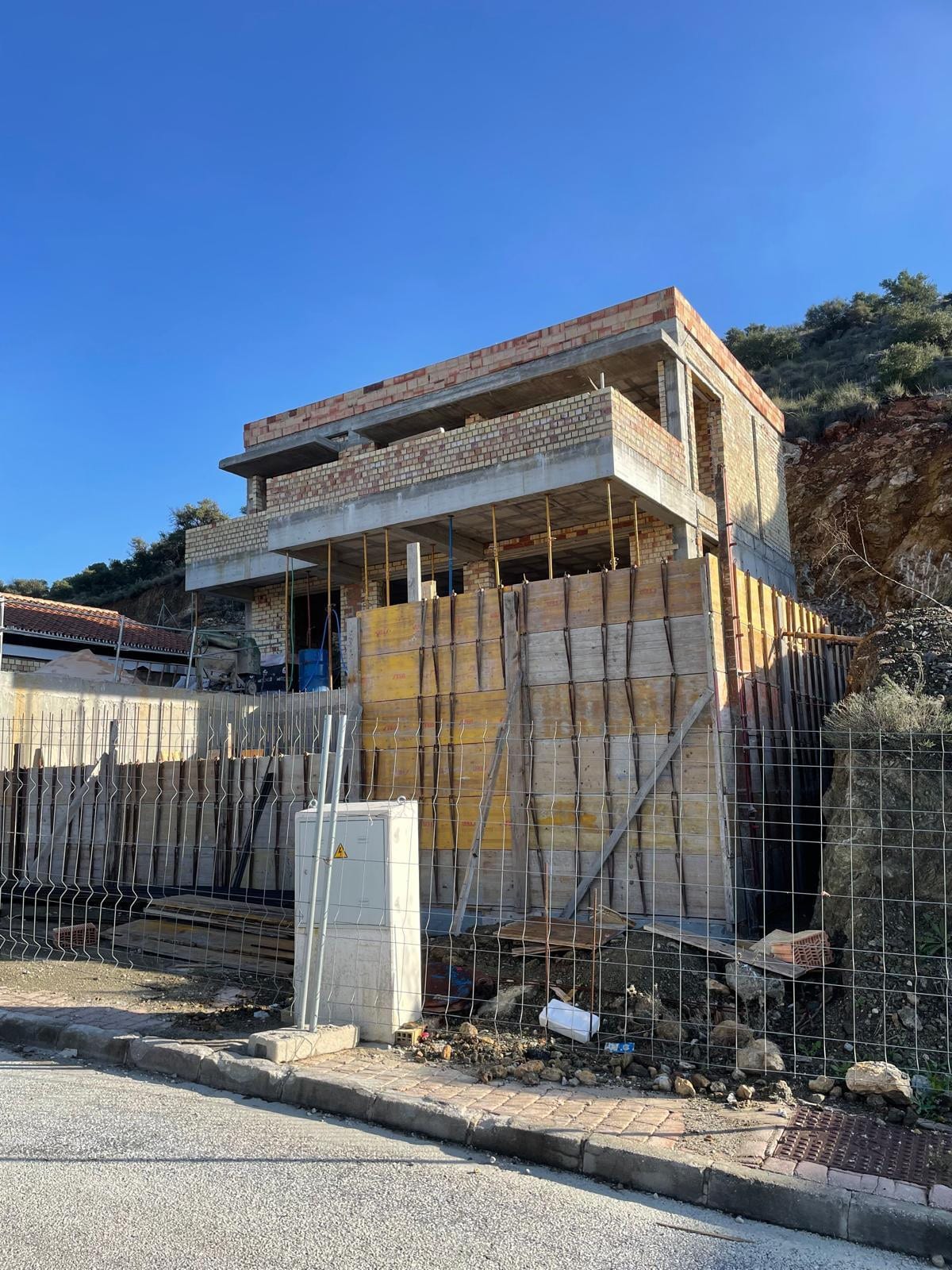 Side view of VILLA ALCALÁ DEL VALLE under construction: a brick side façade across two storeys with concrete overhangs, yellow and timber formwork panels along the façade, metal props, temporary construction fencing and an excavated rocky slope in the background.