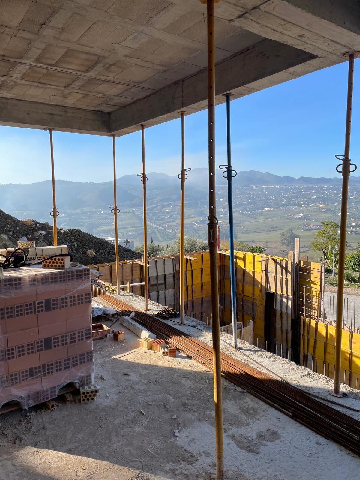 Interior construction view of VILLA ALCALÁ DEL VALLE beneath a concrete floor slab supported by yellow metal props, with stacked bricks and reinforcement bars on the floor; yellow formwork panels and a wide mountain landscape in the background.