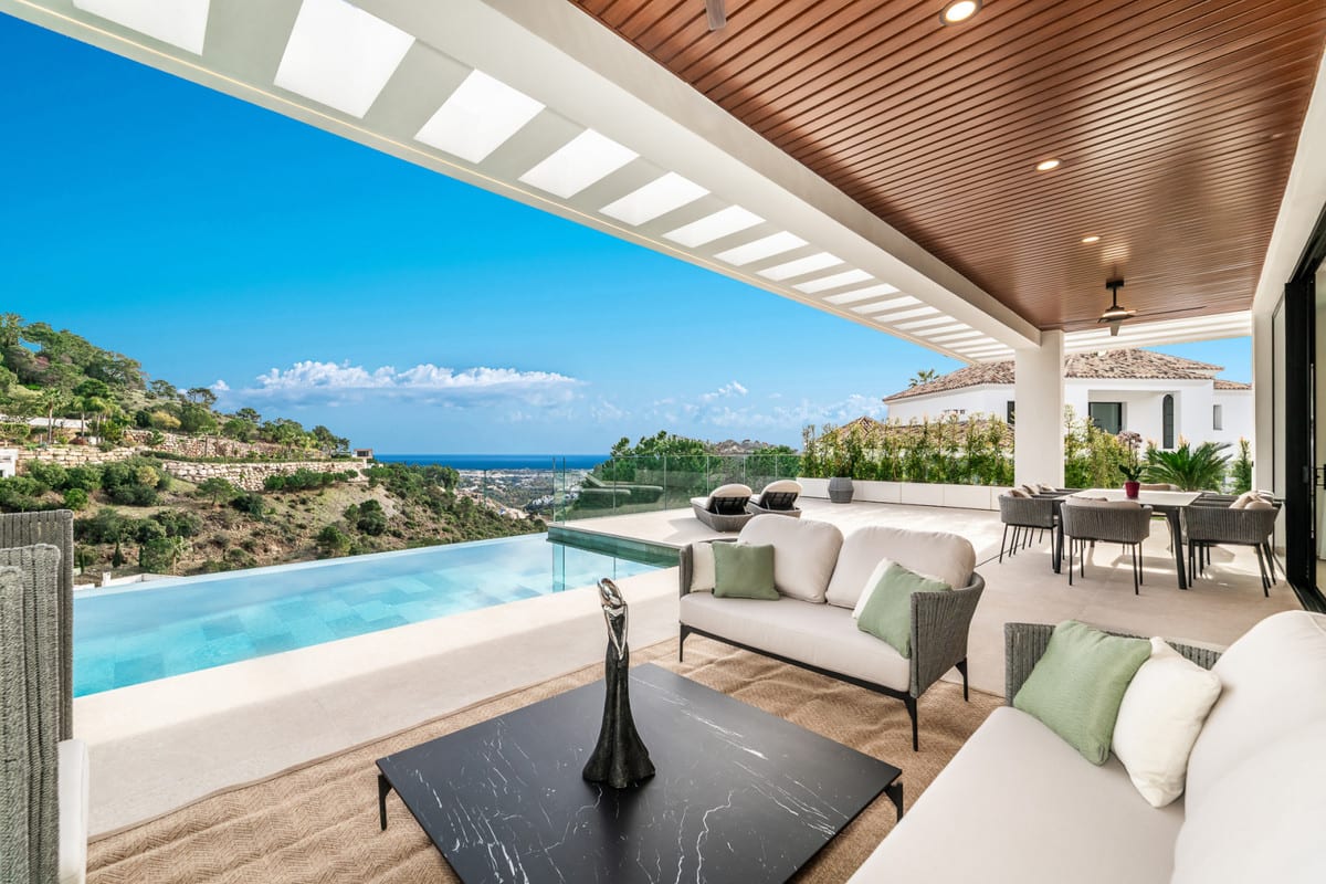 Covered terrace of VILLA OMEGA beneath a timber slatted ceiling, with lounge seating beside the infinity pool and views towards the hills and sea.