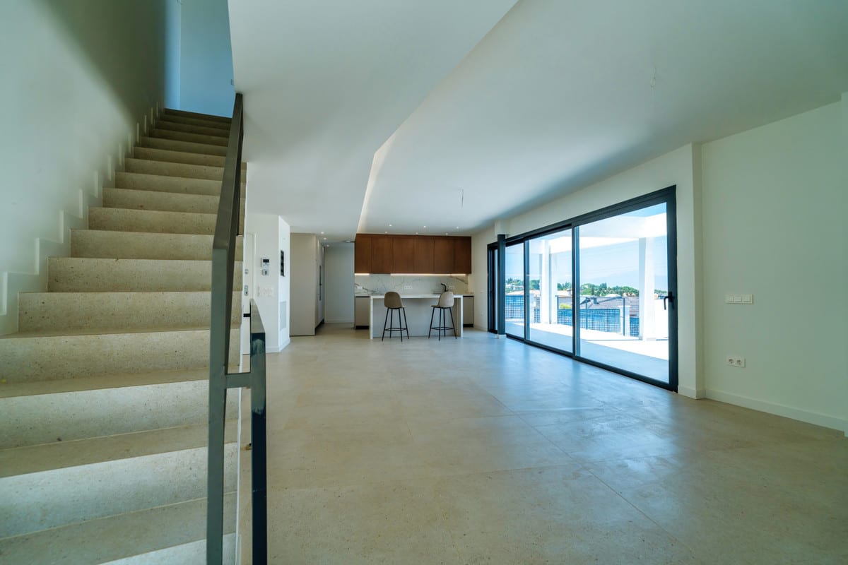 VILLA SANTHAI 3 Y 9 open-plan living space with light tiled flooring, staircase with dark metal balustrade, kitchen in the background and wide sliding doors