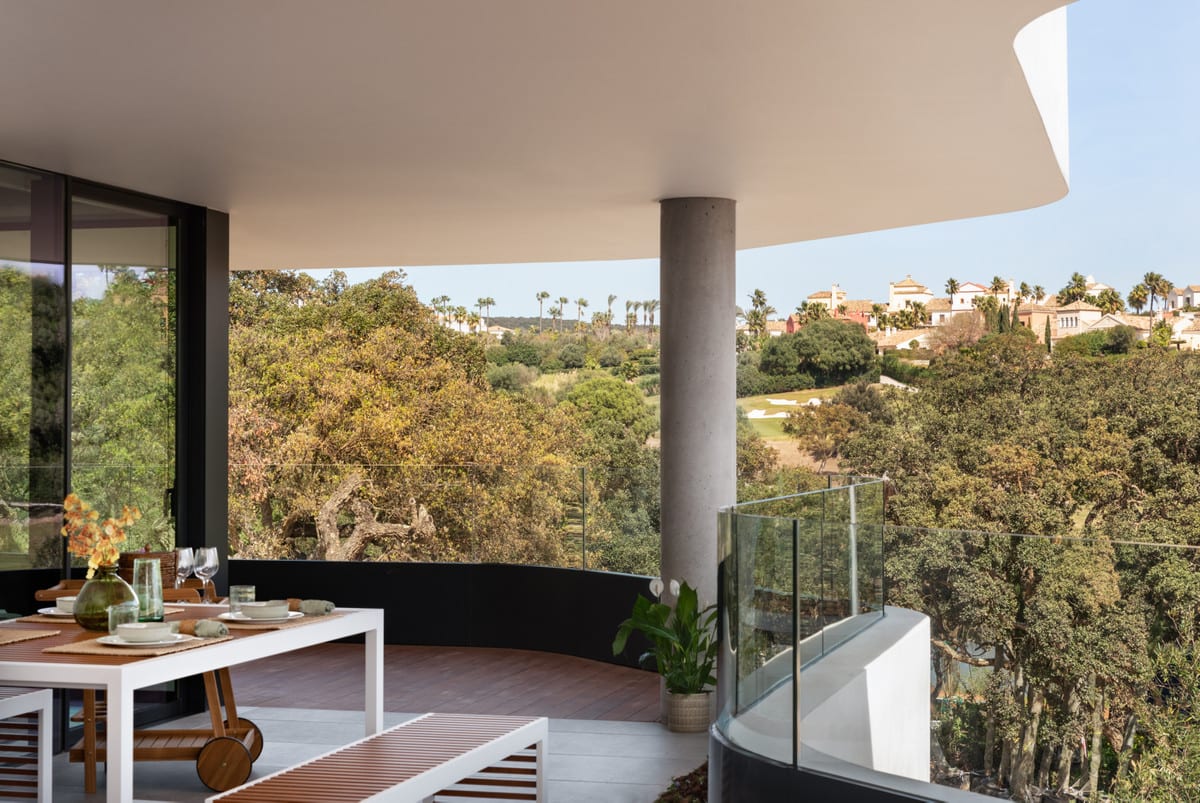 A covered terrace at VILLAGE VERDE with a set dining table, glass balustrade and views of trees and neighbouring homes.