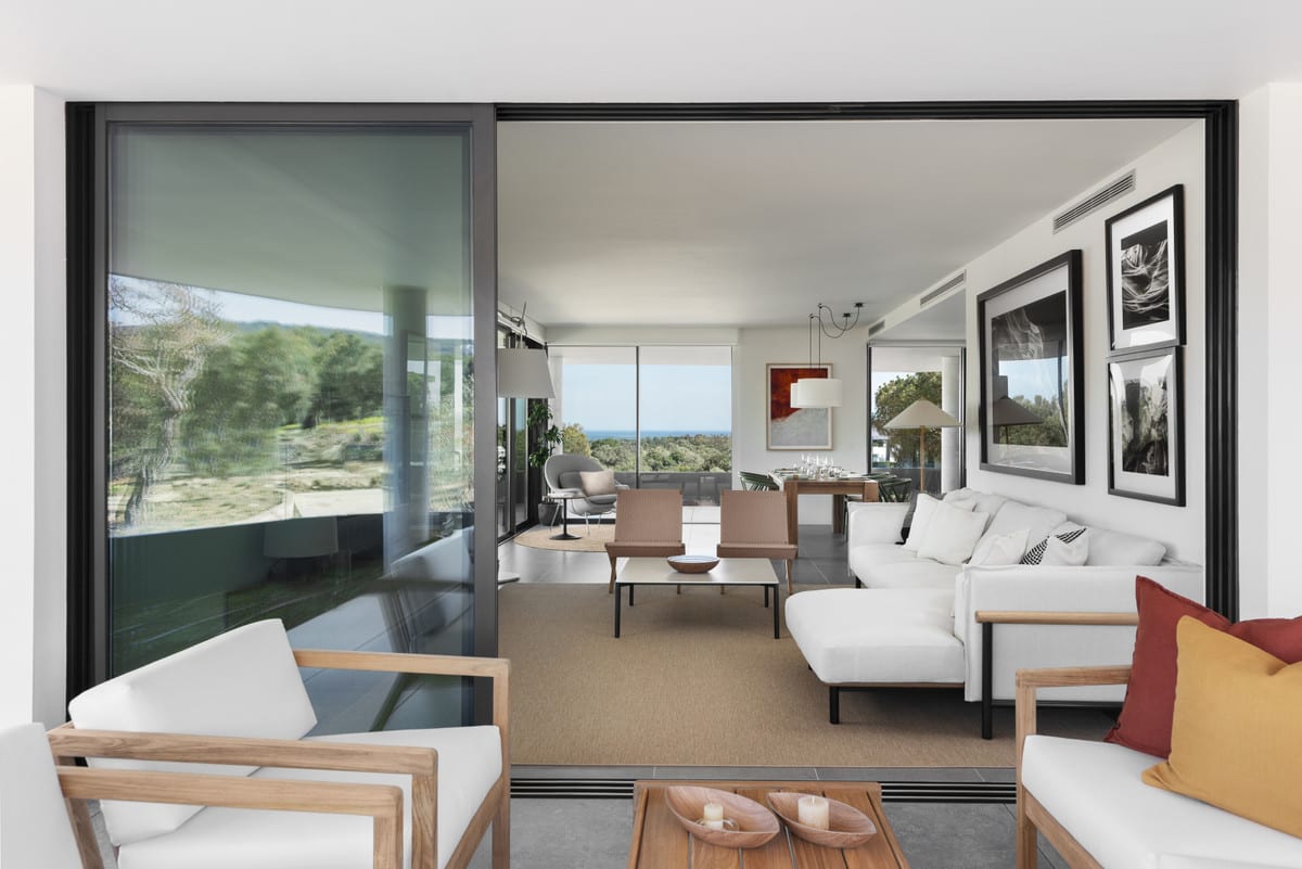 From the terrace seating area, open sliding doors reveal the VILLAGE VERDE living room with a white sofa, timber chairs and framed photographs.