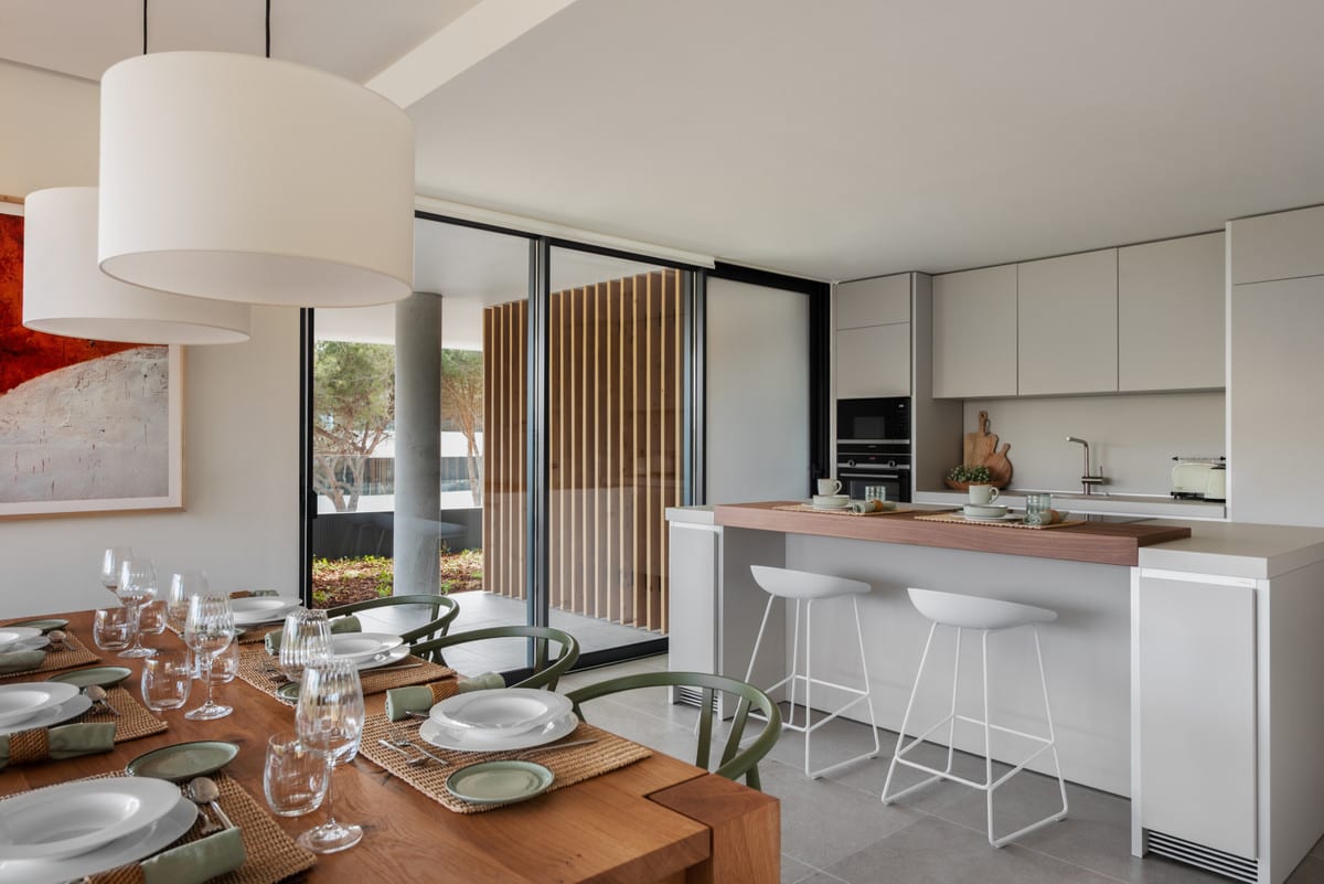 VILLAGE VERDE open-plan kitchen and living area with a laid wooden dining table, white kitchen island with timber worktop and two white bar stools, light-grey kitchen cabinets and glass sliding doors to the terrace