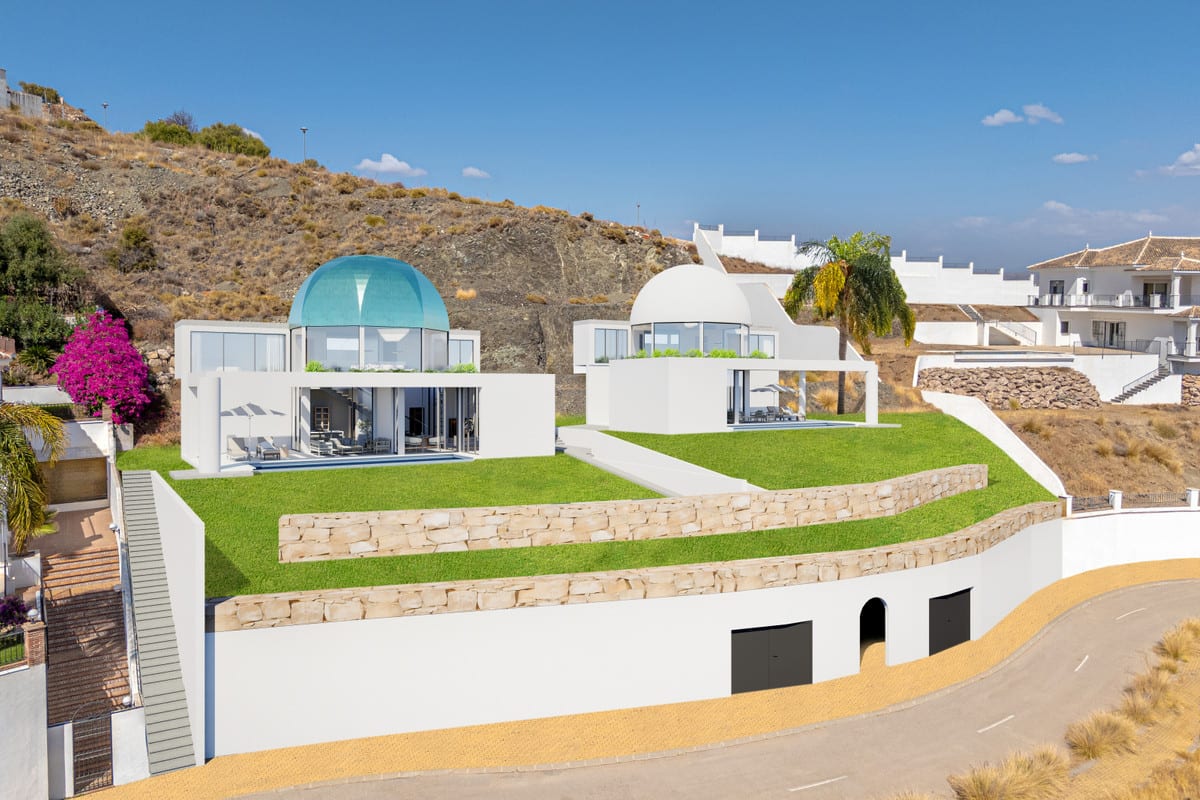 Project visualisation of VISTA DEL REY MIA&AIM from a bird’s-eye view, with two white cubic volumes, a turquoise glass dome and a white dome, glazed façades, tiered lawns, natural-stone retaining walls and garage doors at the base of the building.
