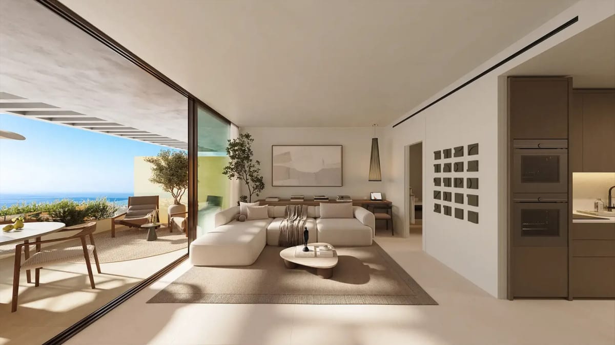 Project visualisation of WATERFALL RESIDENCES with an open-plan living room, cream corner sofa, round coffee table, dark brown kitchen wall with integrated appliances and sliding glass doors to a terrace with a dining area and sea views.