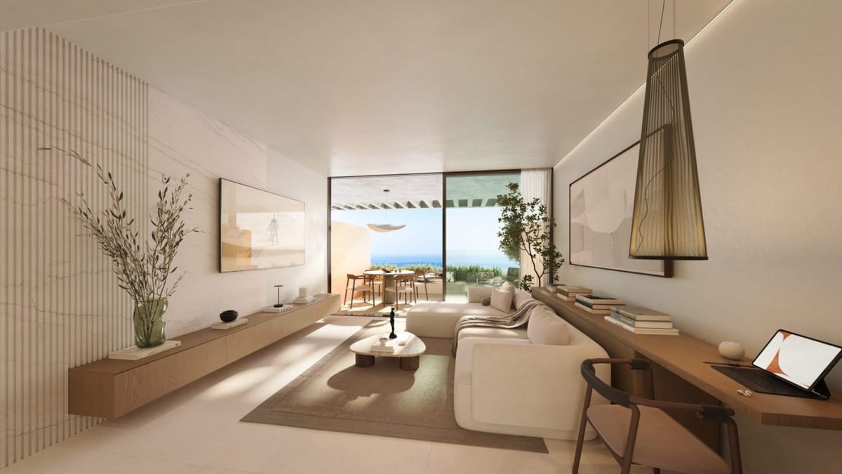 Project visualisation of WATERFALL RESIDENCES with a living room, cream corner sofa, long floating wooden wall shelf, wooden worktop with a chair and tablet, and sliding glass doors to a terrace with the sea in the background.