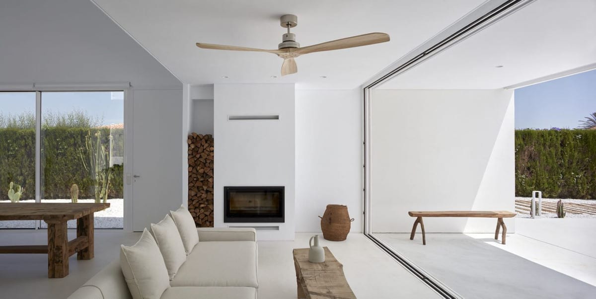 WHITE interior with cream sofa, wooden ceiling fan, fireplace, log storage and fully opened sliding glass wall.