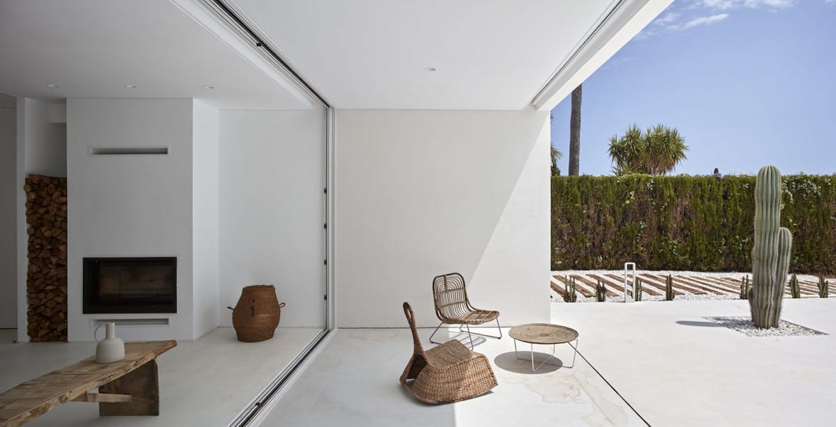 WHITE with open white outdoor area, sliding glass wall, a rattan chair, round table and cactus in gravel.