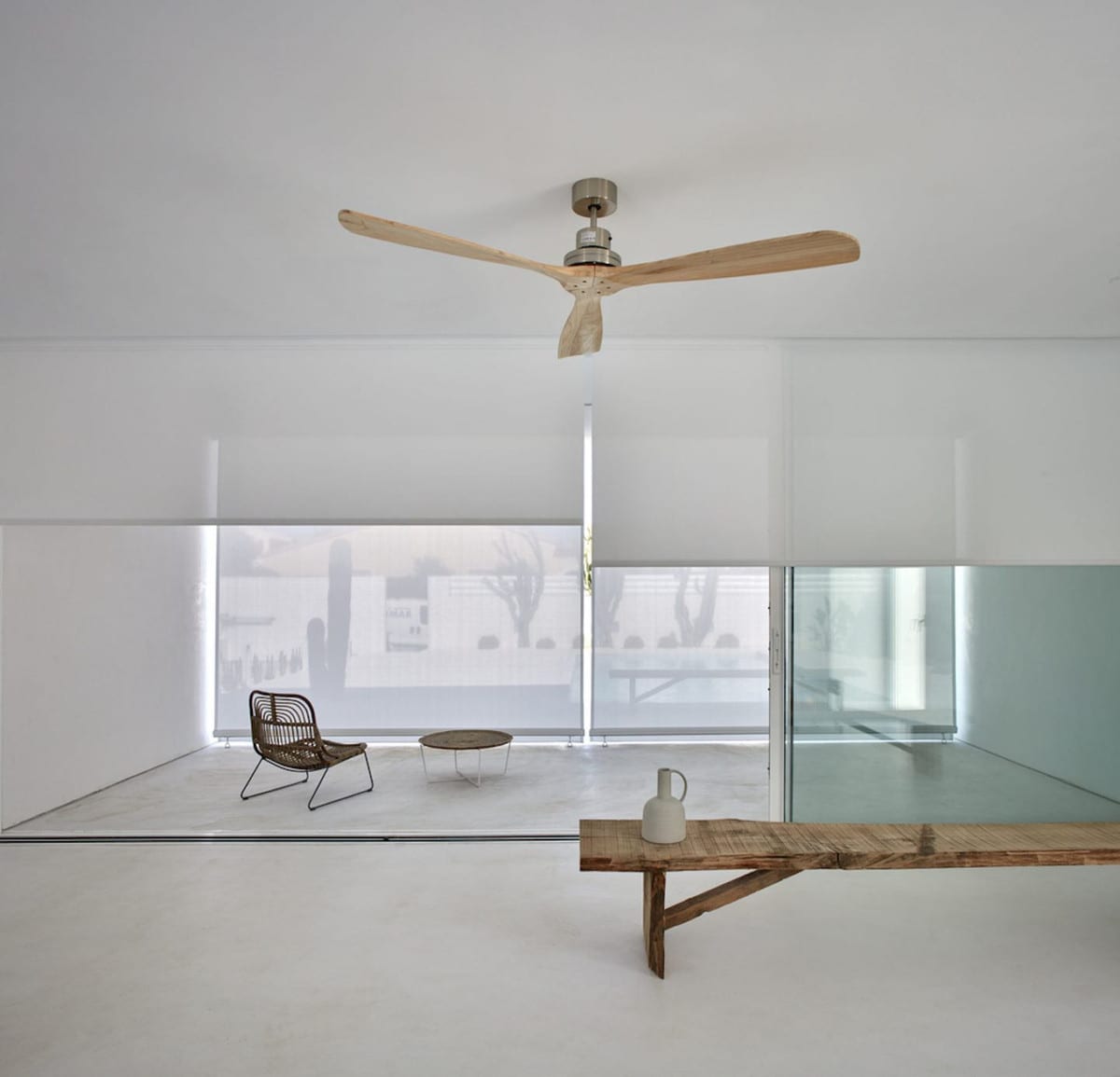 WHITE interior with wooden ceiling fan, white blinds, glass wall, rattan chair and wooden bench with ceramic jug.