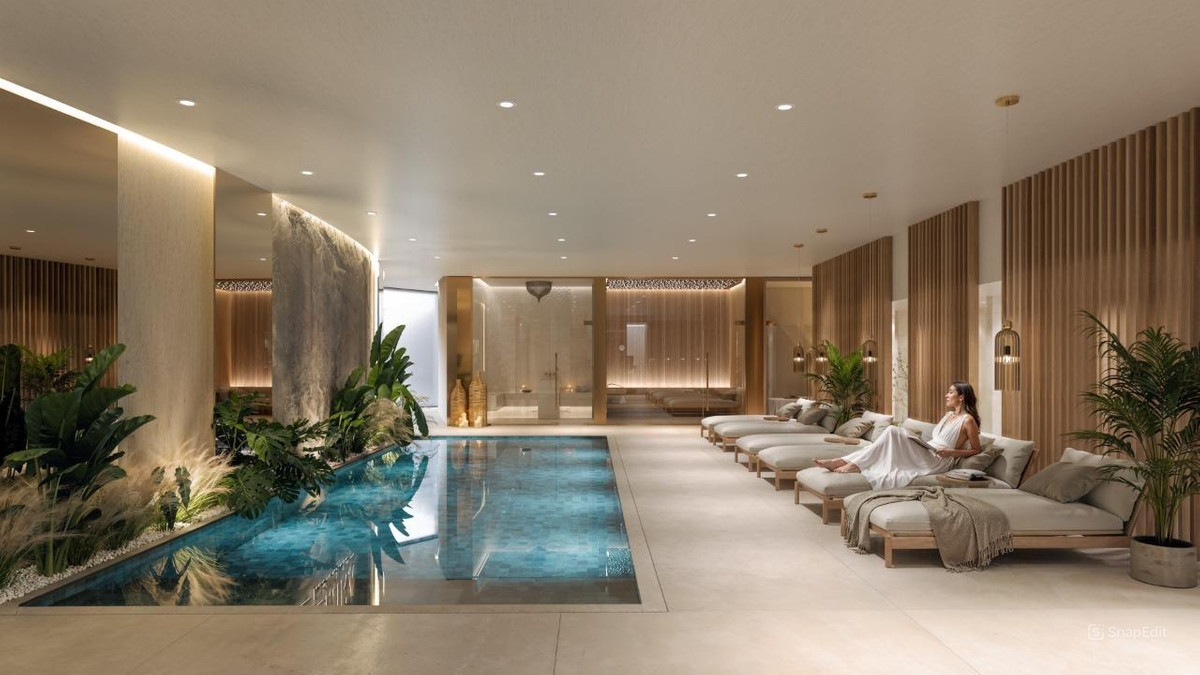Visualisering af en aflang indendørs pool med planter, oplyste søjler, trælameller på væggene og en række liggestole.