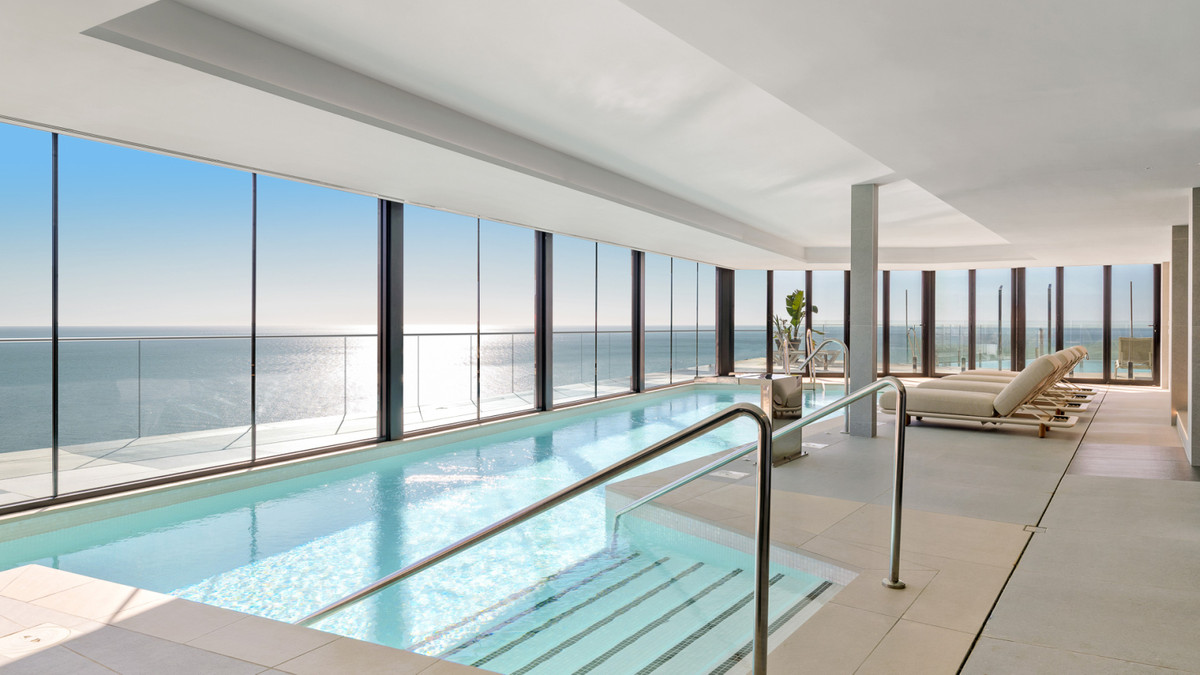 Aflang indendørs pool langs vinduer fra gulv til loft, med siddepladser ved bassinet og udsigt over havet.