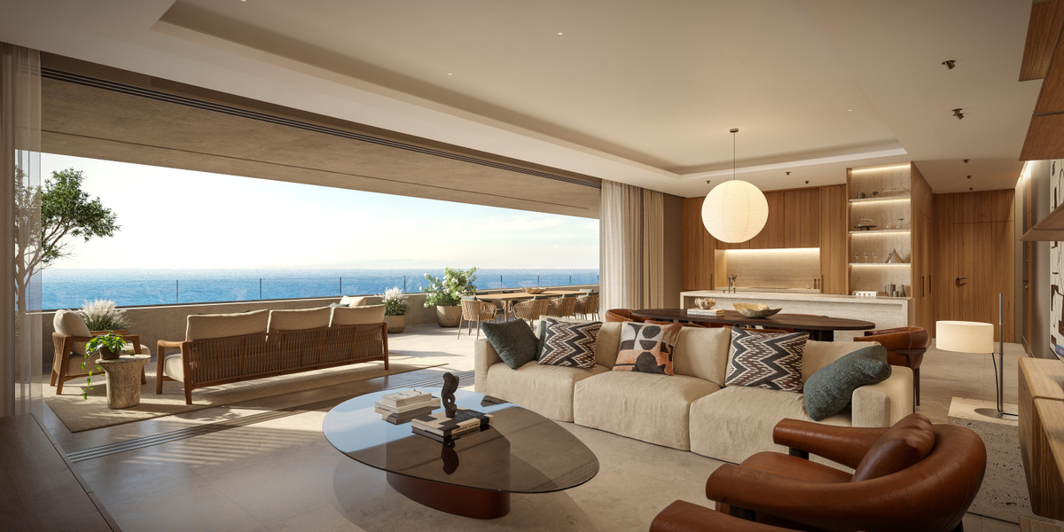 Visualisering af et opholdsrum med lys sofa, rundt sofabord og træbeklædning; en bred åbning forbinder siddeområdet med en terrasse og havet i baggrunden.